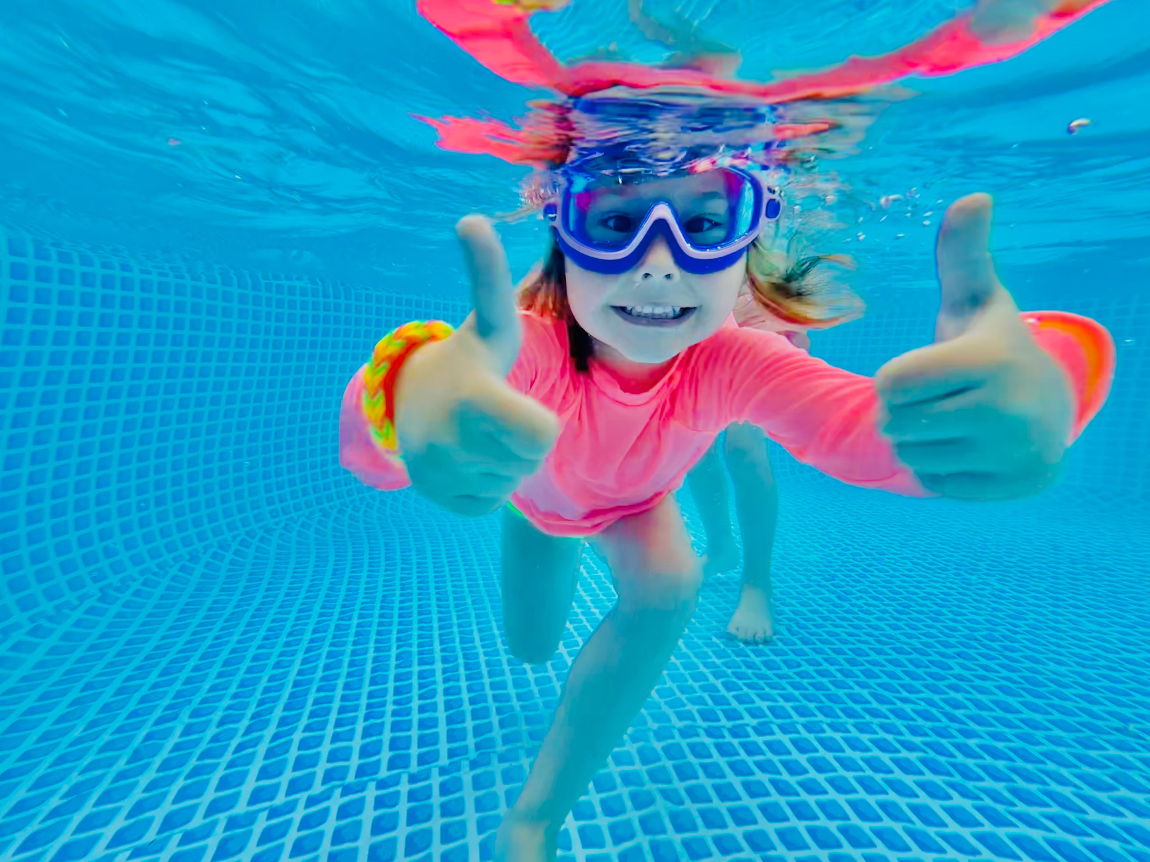 Girl swimming in pool under water wearing a neon coral rash guard with purple goggles giving a thumbs up.